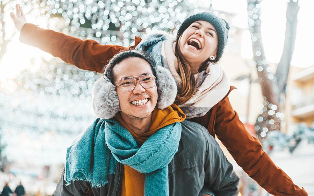 A man gives a woman a piggyback ride outdoors in winter attire near Sterling Ranch CO. Both are smiling and laughing, with festive lights blurred in the background.