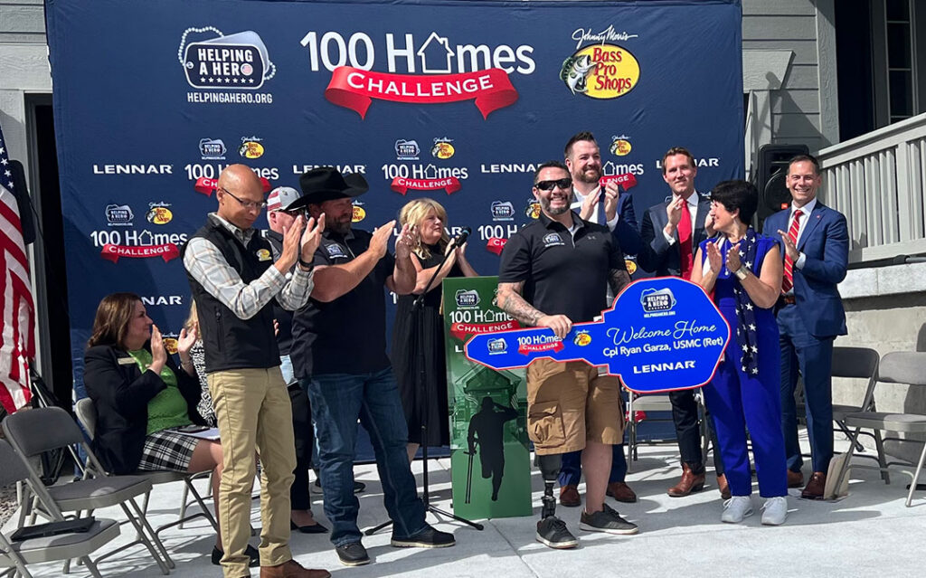 A group of people stand on a stage, applauding as a man in sunglasses holds a large ceremonial key in front of a “100 Homes Challenge” backdrop at Sterling Ranch Colorado, southwest of Denver.