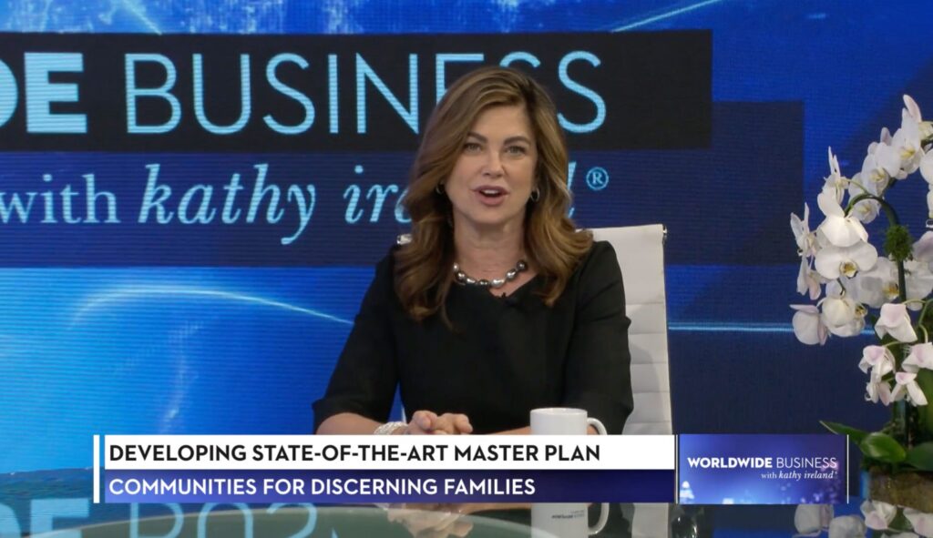 A woman sits at a news desk with a mug and flowers, speaking during a segment titled “Developing State-Of-The-Art Master Plan” on “Worldwide Business with kathy ireland,” highlighting innovative community planning in Sterling Ranch Colorado near Littleton CO in the Denver metro area.