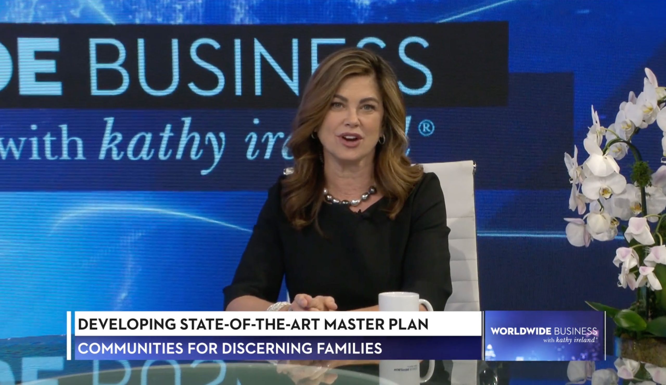 A woman sits at a news desk with a mug and flowers, speaking during a segment titled “Developing State-Of-The-Art Master Plan” on “Worldwide Business with kathy ireland,” highlighting innovative community planning in Sterling Ranch Colorado near Littleton CO in the Denver metro area.