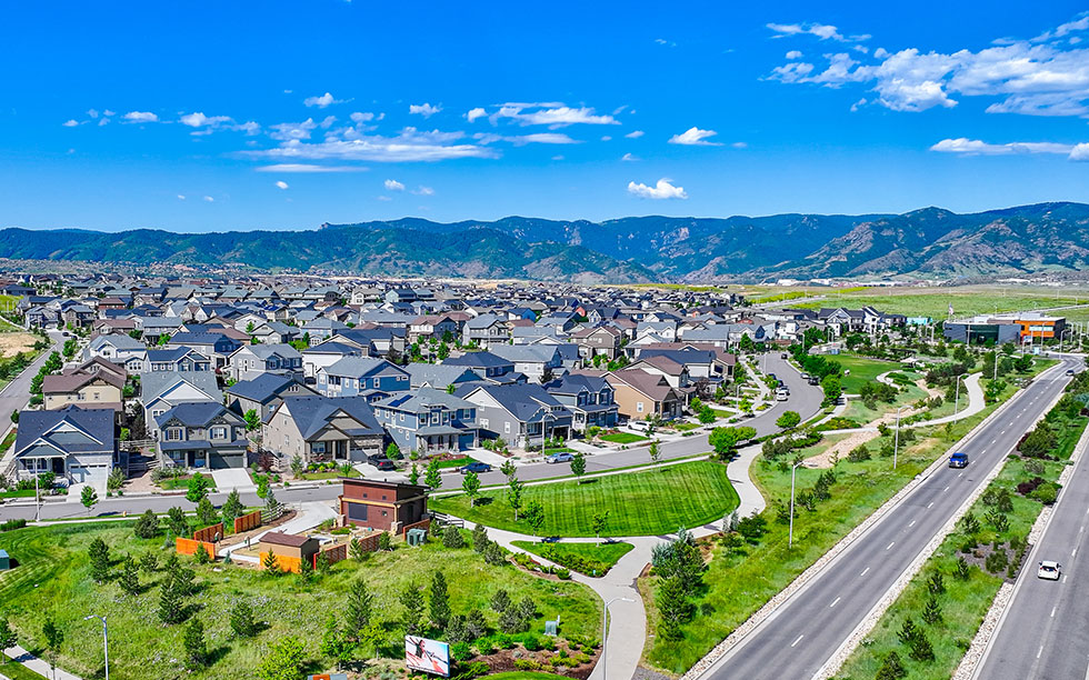 Aerial view of Sterling Ranch Colorado, a suburban neighborhood in Littleton CO with houses, green lawns, winding roads, and nearby mountains under a clear blue sky.