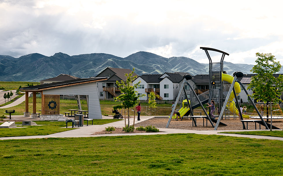 Playground with slides and a climbing structure in a park, with picnic tables, people, and mountains and apartment buildings in the background—located in the Sterling Ranch community, southwest of Denver in the Denver metro area.