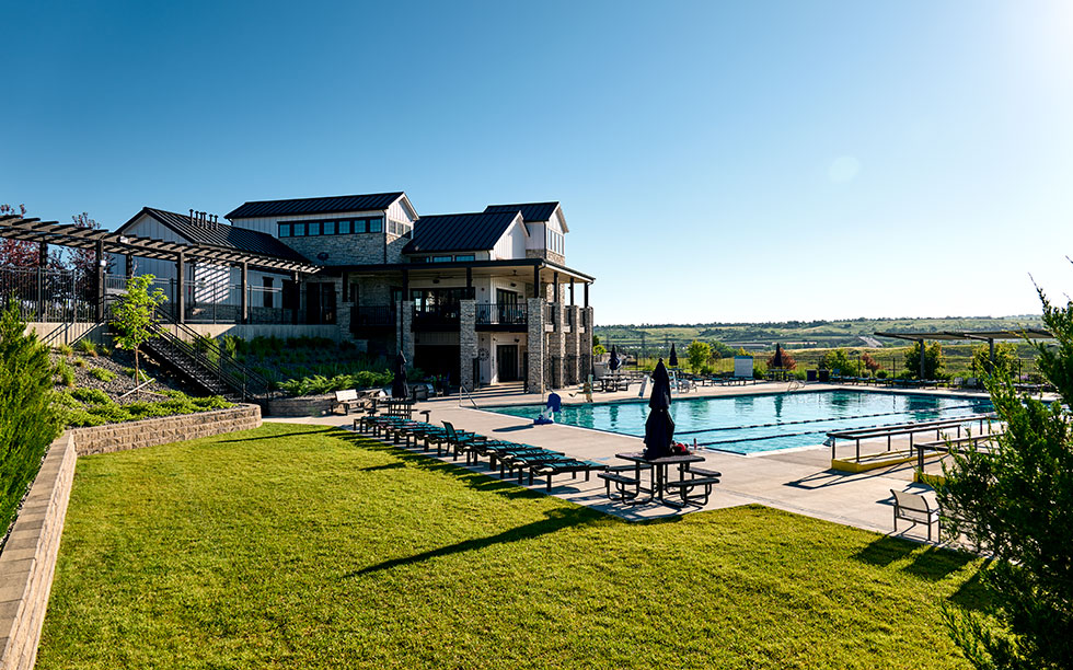Modern clubhouse with a large outdoor swimming pool, lounge chairs, umbrellas, and well-maintained green lawn under a clear blue sky—located in the heart of Sterling Ranch, southwest of Denver in Littleton, CO.