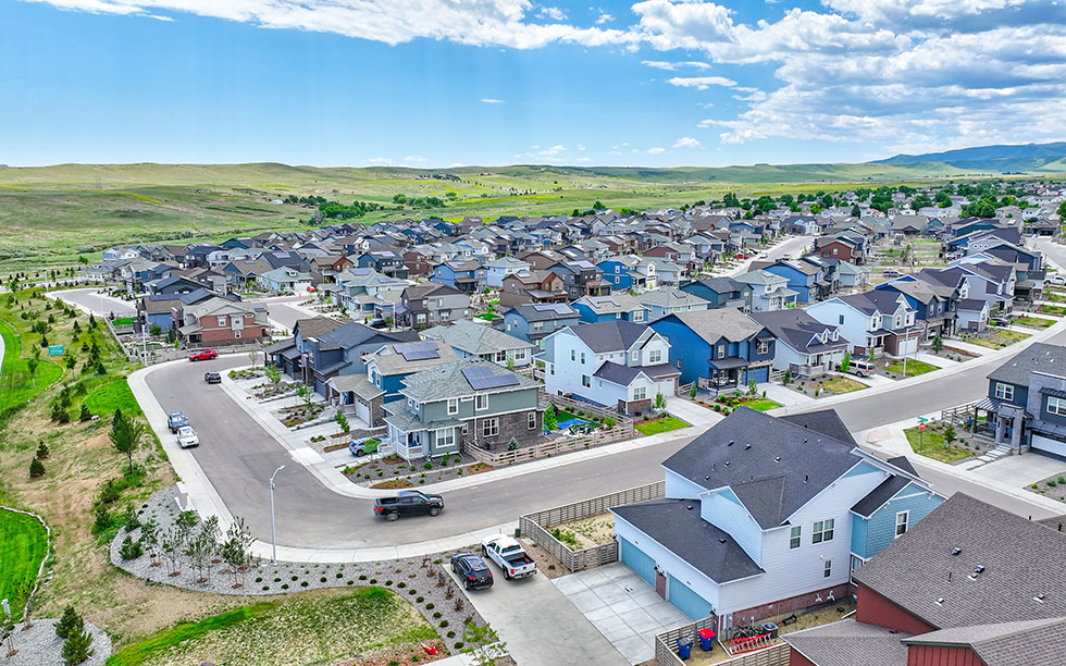 Aerial view of Sterling Ranch CO, a suburban neighborhood southwest of Denver, with rows of houses, green lawns, curved streets, and open fields in the background under a partly cloudy sky.
