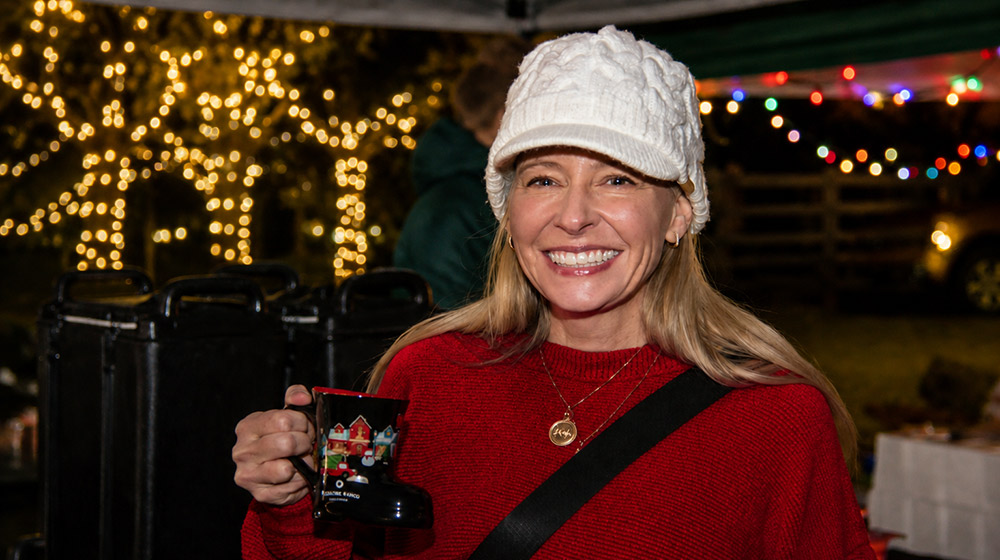 Woman in a white knit hat and red sweater smiles while holding a mug at an outdoor event with string lights in the background.